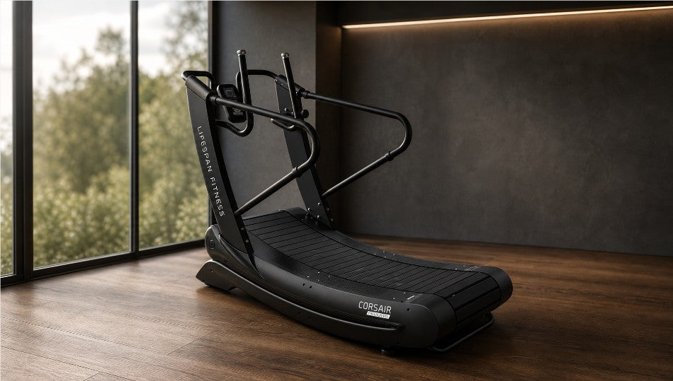 A black Cosair elliptical trainer with dual handles and a curved running deck, positioned in a modern gym setting with a dark textured wall, wooden floor, and large windows showing green trees outside.