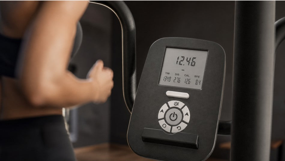 Close-up of a black digital exercise bike console with a display screen and control buttons, with a person's arm blurred in the background on a gym machine.