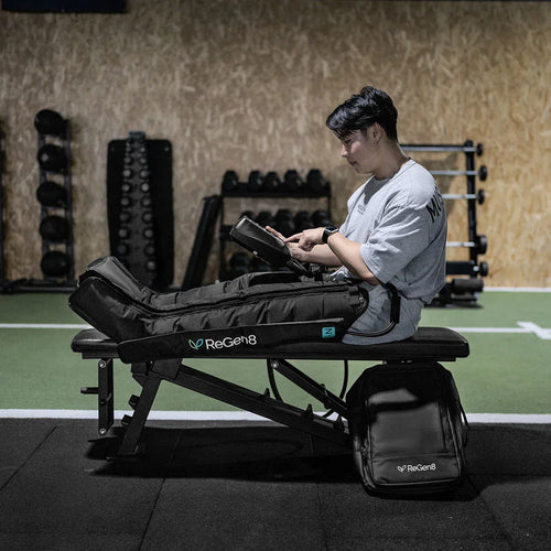 Person using ReGen8 Compression Recovery Boots while reading on a bench in a gym setting.